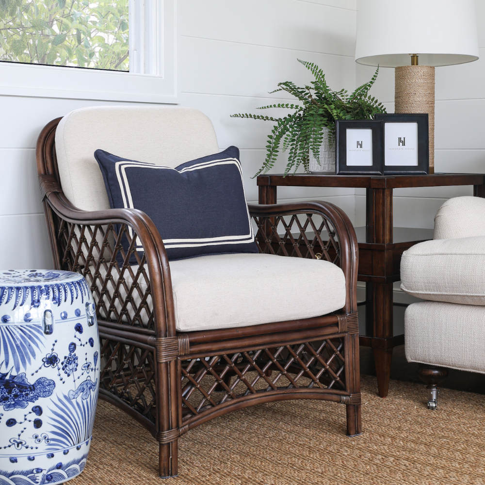 Rattan arm chair with white cushion and navy pillow in a room with a table, lamp, and plant.