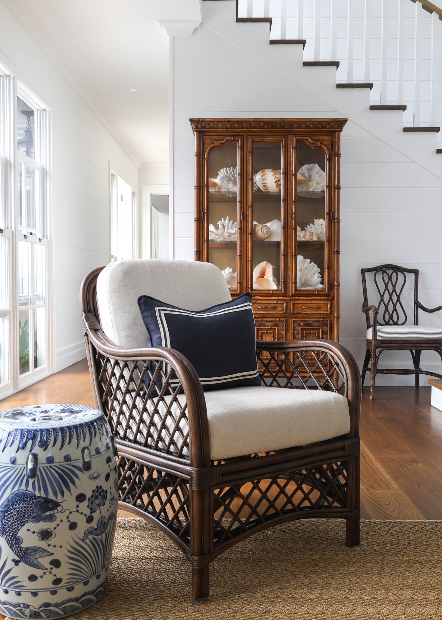 Rattan chair with white cushion and navy pillow in a room with wooden floor and staircase.